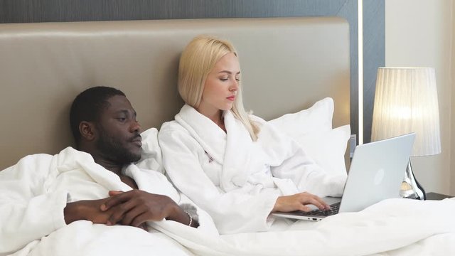 African man and caucasian woman with laptop in hotel, they lie on bed together, discuss where to go during their travel. suitcase near them.
