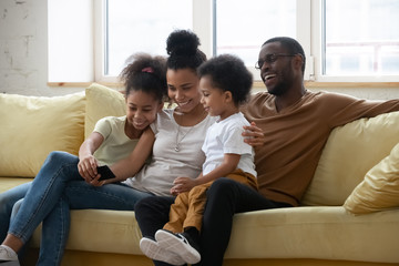 Mommy, daddy, look here. Happy african kid girl sitting on couch at home holding cellphone showing cute video to little preschool brother and smiling parents mom and dad, making call, using mobile app