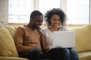 Fototapeta premium Happy black couple speak with friends online making video call. Smiling mixed race married man and woman sitting on coach in living room use laptop for distance conference calling