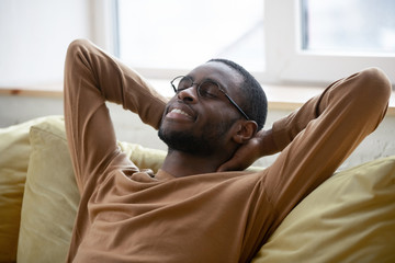 Mental rest. Calm peaceful young african man relaxing on couch at home sitting in comfortable pose feeling delight and pleasure, millennial black guy resting from work with closed eyes and happy smile