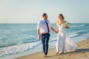 bride and groom walk on beach happy and laughing by the seashore.