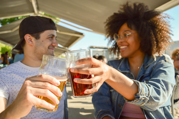Young beautiful happy Caucasian and Afro couple of friends toasting with glasses of beers outside. Smiling while cheering together. Concept about people, lifestyle and holiday. 