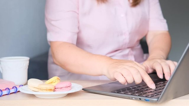 fat woman sits at a computer and surfs the Internet, next to a laptop there is plate with macaroon pastries, concept of diet, high-calorie nutrition, weight loss problems, body positive