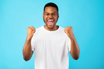 Fototapeta premium Emotional Black Guy Shouting Yes Shaking Fists On Blue Background