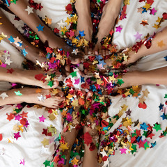 Group of 5 women sit in circle in bed close up top view well-groomed female barefoot feet legs covered with shiny multi colored confetti pieces star shaped, hen party celebration, spa skincare concept