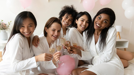 Horizontal view five multi racial girls friends in white bath robes holding champagne glasses smile...
