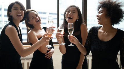Happy multiethnic girls wearing elegant little black evening or cocktail dresses toasting clinking...