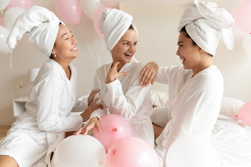 Seated on bed three cheerful multi ethnic women wearing morning gowns towel on heads laughing enjoy spa procedures beauty grooming treatments, friends intimates celebrating bachelorette party concept