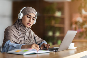 Girl in headscarf having online lesson on laptop at cafe - obrazy, fototapety, plakaty