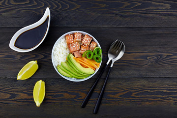 Hawaiian salmon fish poke bowl with rice, avocado, cucumber, carrot on dark rustic background.