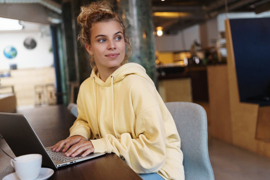 Smiling Woman Sitting In Cafe With Laptop, Looking Outside Window Pleased. Creative Female Freelancer Getting Inspiration Working Outdoors In Coffee Shop. Girl Studying Online In Cafe With Laptop