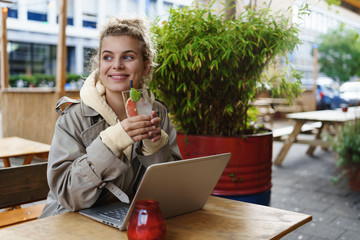 Dreamy happy girl enjoying drink in cafe while having break work. Smiling woman working remote in coffee shop with laptop computer, drinking cocktail. Student studying online on fresh air in cafe