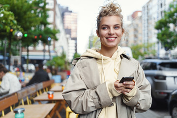 Happy beautiful girl waiting for someone outside cafe, holding mobile phone and smiling. Pretty woman order taxi in smartphone application, standing on street. Female using carsharing to get work