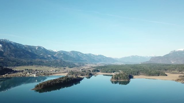A drone flight above the Faaker See in Austria. There are high Alpine ranges around, reflecting in the crystal clear and calm surface of the lake. Small villages around the lake. Snow capped mountains