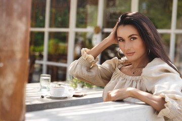 Sensual brunette woman with dreamy smile, turn back looking camera as sit outdoor cafe at summer sunny day, enjoying nice cup of coffee favorite coffeeshop, eating lunch. Sexy female at restaurant