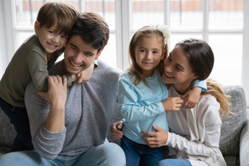 Happy adorable small children cuddling affectionate loving parents, relaxing together on comfortable sofa. Smiling young couple enjoying spending time with preschool cute kids siblings, family pastime
