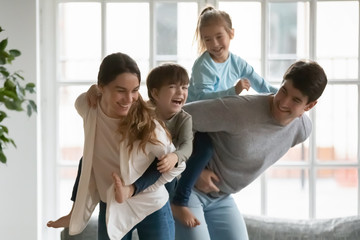 Playful young mixed race mother holding on back emotional little kid son while smiling father giving piggyback ride to happy preschool daughter, parents playing with energetic children at home.