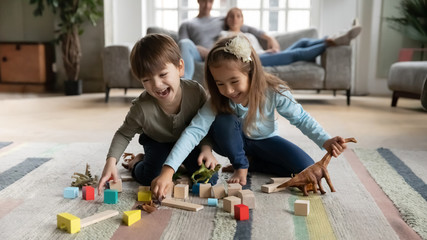 Overjoyed funny little children siblings playing toys on floor carpet while parents resting on...