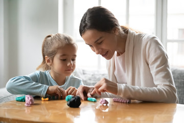 Head shot smiling pretty young mixed race nanny teaching little funny preschool kid girl making figures with colorful playdough, sitting together at table, happy bonding family weekend activity.