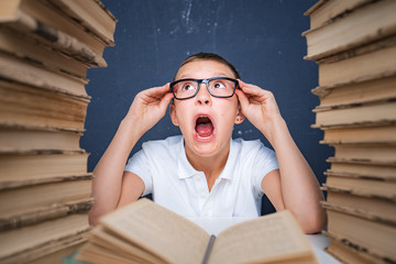 Smart boy in glasses getting crazy of reading book while sitting between two piles of books.