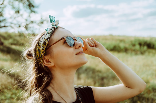 Close up of a young woman in the field with sun glasses reflecting beautiful clouds, boho style