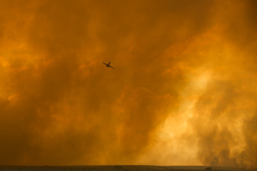Orange hanging curtains of smoke from a large wildfire partially obscuring a firefighting plane in the desert