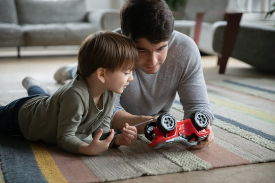 Caring young father lying on floor carpet with little adorable kid son, teaching fixing toy car in living room. Interested small child boy involved in repairing process with daddy, skills development.