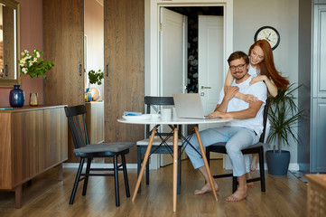 woman tries to cheer up her husband during his work on laptop, man sit with laptop, he is on freelance. redhead lady loves hes husband