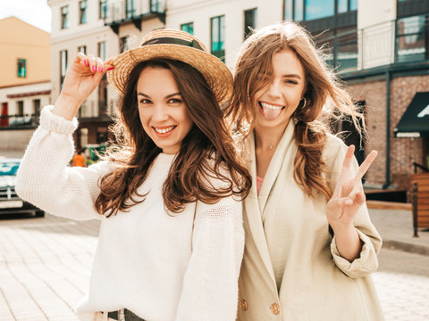 Two young beautiful smiling hipster girls in trendy white sweater and coat. Sexy carefree women posing on street background in hat. Positive models having fun. They show peace sign