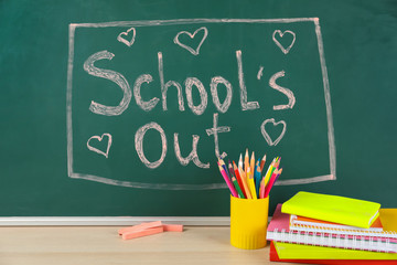 Pieces of color chalk and stationery on table near greenboard with text School's Out. Summer...