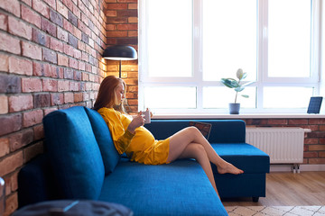 side view on attractive redhead pregnant woman drinking tea at home, woman in yellow dress sits on sofa, have rest