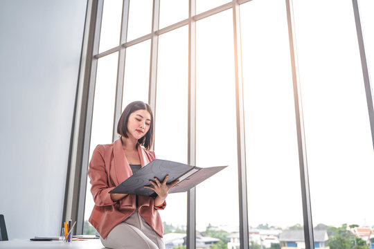 Beautiful Female Asian Worker Holding A Folder File Of Documents And Information On Work Or Business Looking, Reading, Learning From The Documents, While Holding A Pen Working In An Office Environment