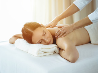 Pretty brunette woman enjoying procedure of back massage in sunny spa salon. Beauty concept. Cold toned picture