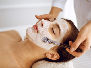 Beautiful brunette woman enjoying applying cosmetic mask with closed eyes. Relaxing treatment in medicine and spa center concepts