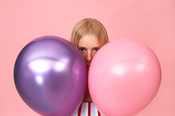Fototapeta na wymiar Isolated shot of mysterious young blonde female with freckles and facial piercing posing against pink background hiding herself behind two shiny metallic helium balloons, staring at camera