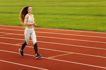 Obraz premium Athletic woman running at the stadium, working out, keep herself in shape, wearing sportswear, white top and pants, recommends doing sports, healthy lifestyle concept