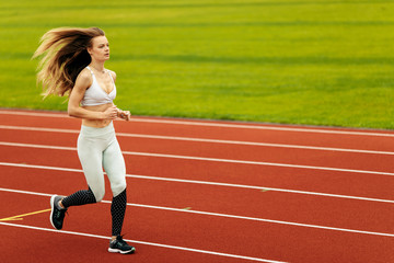 Obraz premium Sporty woman running at the stadium, working out, keep herself in shape, wearing sportswear, white top and pants, recommends doing sports, healthy lifestyle concept