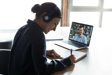 Smiling young indian woman in headphones learning practicing foreign language with confident male tutor distantly on computer. Happy mixed race girl listening to educational webinar, writing notes. - obrazy, fototapety, plakaty