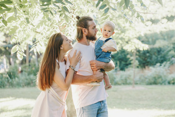 Fototapeta premium Happy smiling mother and father with baby boy in park outdoors. Family Caucasian mom and dad with son walking hugging in park on summer day. Happy family authentic lifestyle concept.