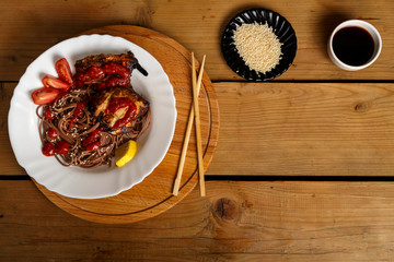 Soba with chicken in a white plate on a round stand with soy sauce and sesame seeds next to...