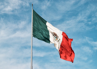 Close-up of a mexican flag flying with the wind on the top of a flagpole with a blue sky and clouds as background.