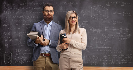 Male and female teacher holding books in front of a blackboard