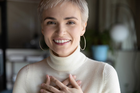 Close up head phot profile portrait of kind young businesswoman holding folded hands on chest, feeling thankful. in modern office. Smiling compassionate female employee involved in charity activity.