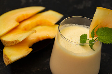 Melon milk shake on a black background. Ripe sliced melon is lying nearby. The smoothie glass is decorated with a sprig of mint and melon crumbs. Summer healthy food concept
