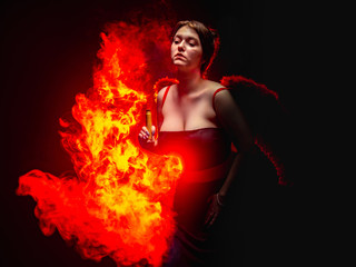 Obraz na płótnie Canvas Devil. Girl with e-cig on the fire background. A woman in a devil costume. A young woman depicts a devil with a vape device. Red wings behind the dancer's back. Witch with fire. Devilish fire.