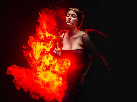 Devil. Girl with e-cig on the fire background. A woman in a devil costume. A young woman depicts a devil with a vape device. Red wings behind the dancer's back. Witch with fire. Devilish fire. © Grispb