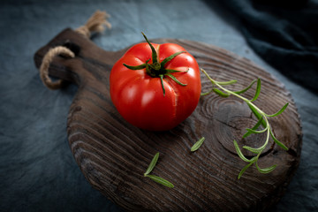 Ripe organic tomato and rosemary sprig on an old chopping board. Ripe vegetable was cracked with juice. Selective focus on the crack.
