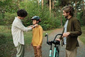 Young careful female helping her son with tying up belts of protective helmet