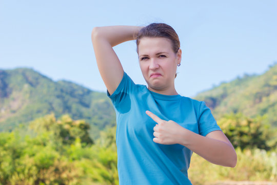 Young beautiful woman, sweating girl with wet spot on T-shirt because of sweat smelly armpits looking at camera, show, point at her armpit with finger. Using deodorant antiperspirant
