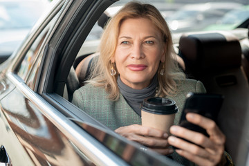 Mature woman having coffee and scrolling in smartphone while sitting on backseat